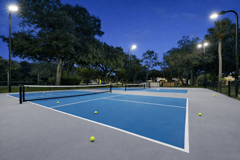 Tennis court with blue surface and white lines, surrounded by trees and street lights.